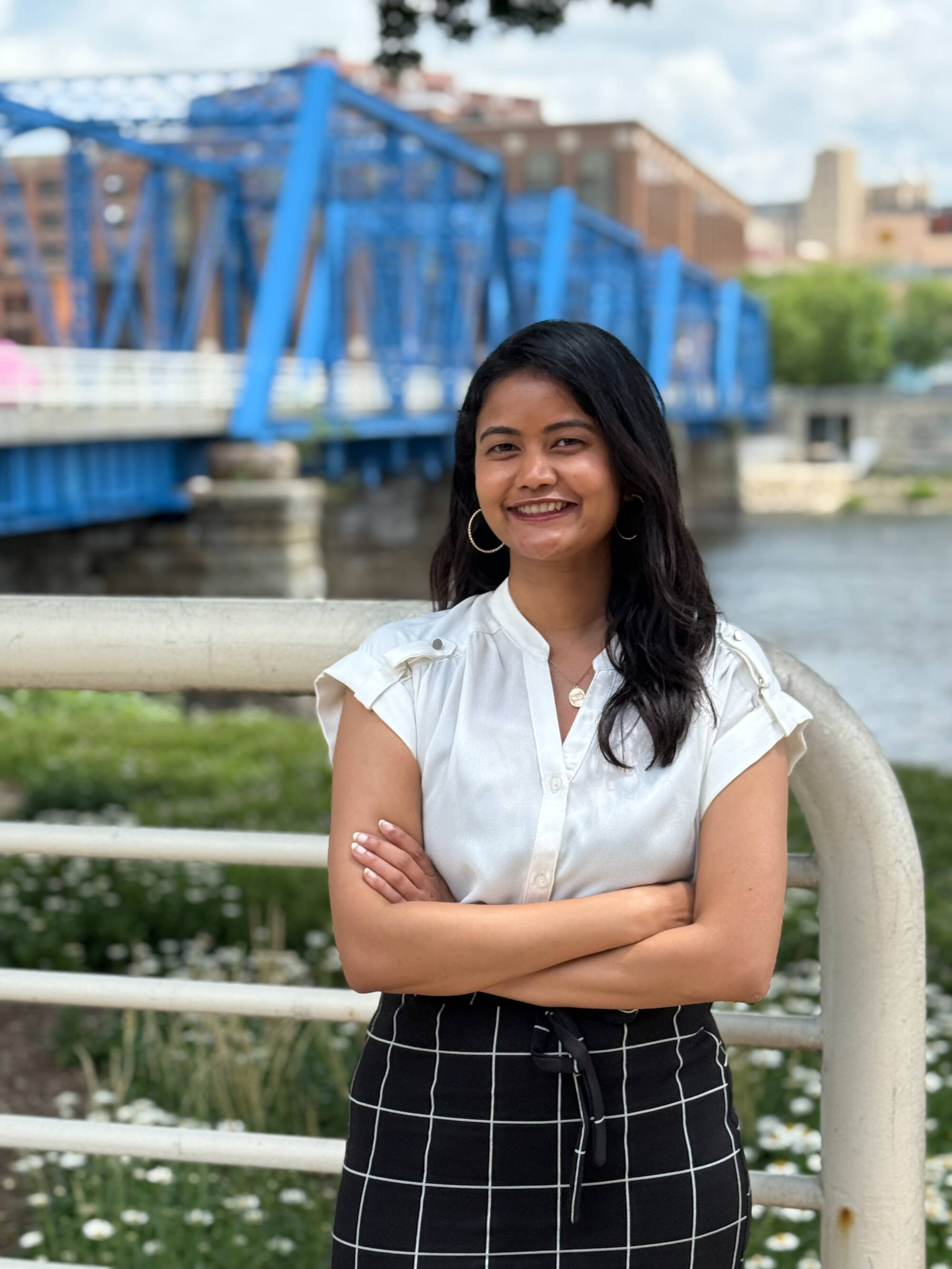 A lady with long dark hair is standing outdoors in front of a blue metal bridge, with her arms crossed and dressed in a white short-sleeve blouse and a black skirt with a white grid pattern. The background includes green grass, flowers, and city buildings